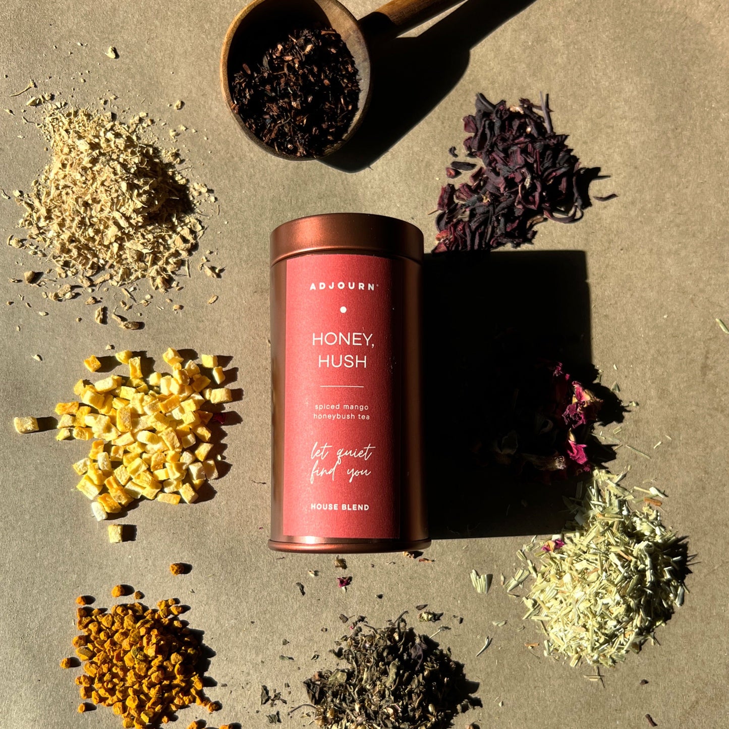A tin of Adjourn Honey, Hush honeybush Blend herbal tea is displayed on a textured beige surface, surrounded by loose tea ingredients in a wooden spoon and scattered piles, with a wooden spoon scoop of the tea above.