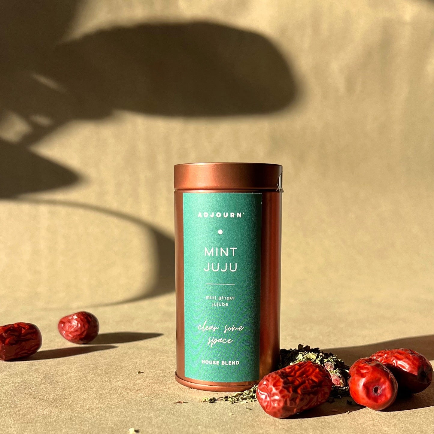 A cylindrical metal tin of AJOURN Mint Juju loose leaf tea stands on a beige surface with dried red jujubes and loose tea scattered around it. The background is a matching beige paper backdrop with dramatic leaf shadows.