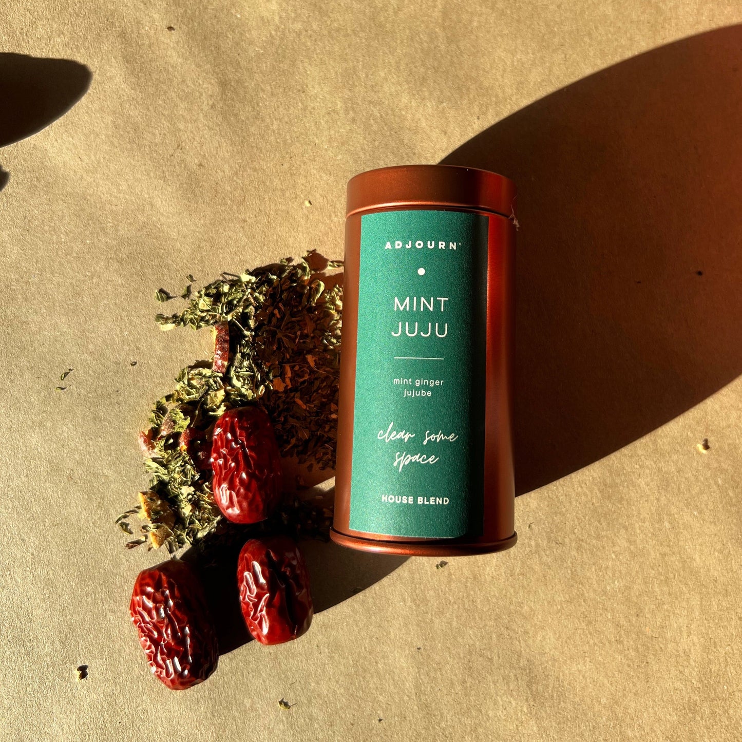 A cylindrical tin of ADJOURN Mint Juju loose spice blend sits on a textured beige surface, styled with dried mint leaves and red dates under warm, direct sunlight.