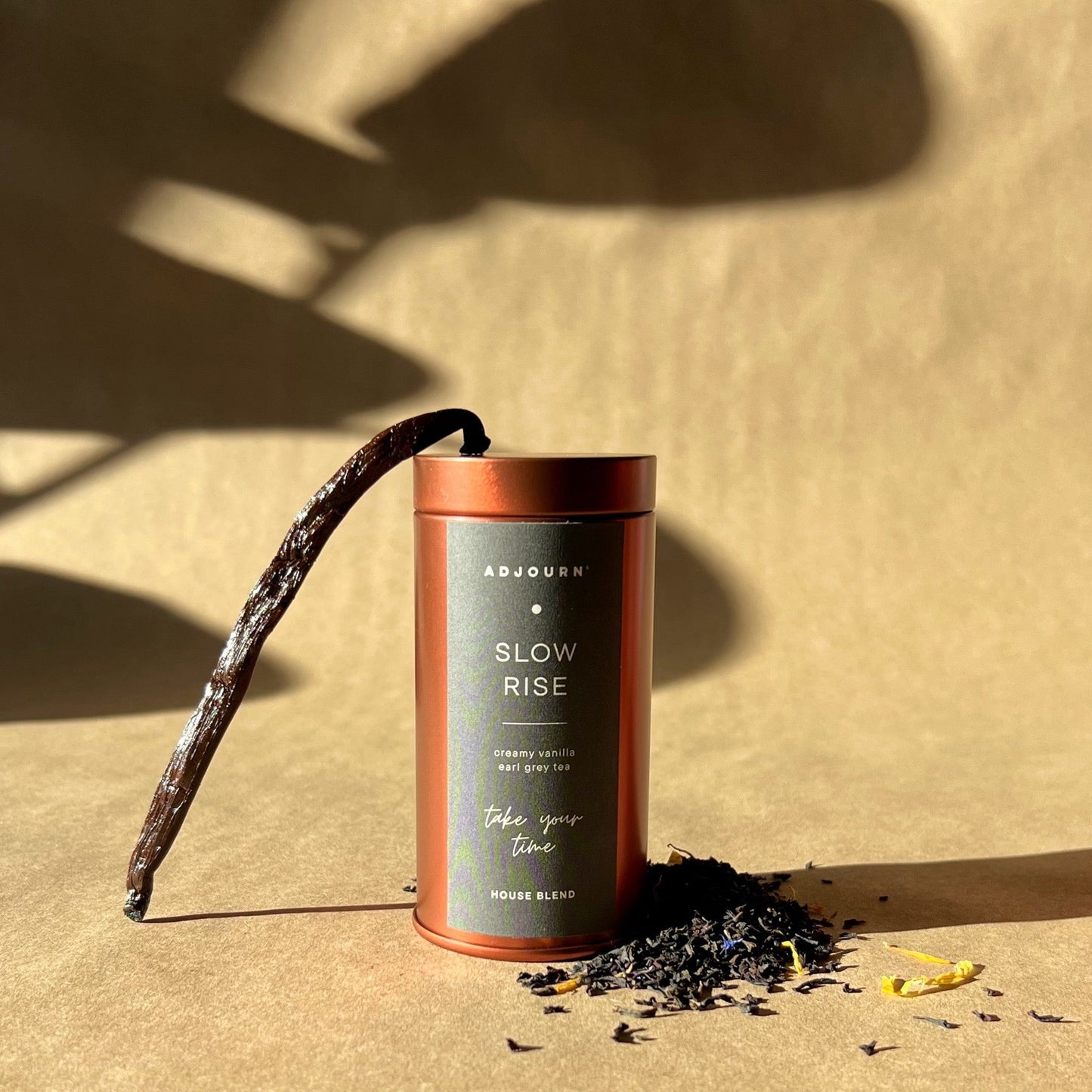 A cylindrical metal tea tin labeled 'SLOW RISE' stands on a warm beige surface with loose tea leaves scattered around it and a long dried vanilla bean leaning against the tin. Strong directional sunlight casts dramatic shadows on the background.