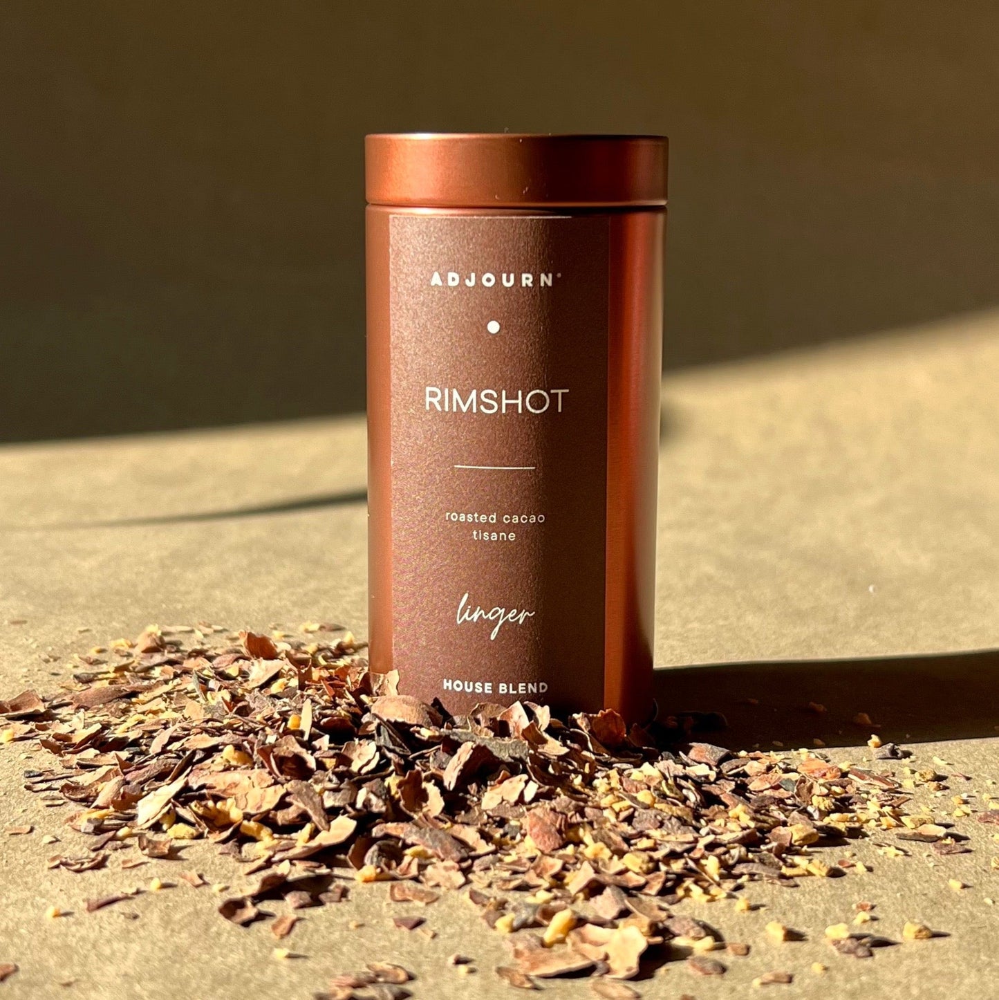 A cylindrical copper-toned metal tin of Adjourn Rimshot loose cocoa beans stands on a textured surface with scattered cocoa nibs in the foreground, lit by warm directional sunlight against a neutral background.